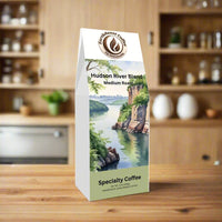 Front view of EarthRoast Coffee's Hudson River Blend, a medium roast specialty coffee. Features a scenic illustration of the Hudson River with cliffs, lush greenery, and a city skyline in the distance, emphasizing its organic, sustainably sourced quality and balanced flavors.